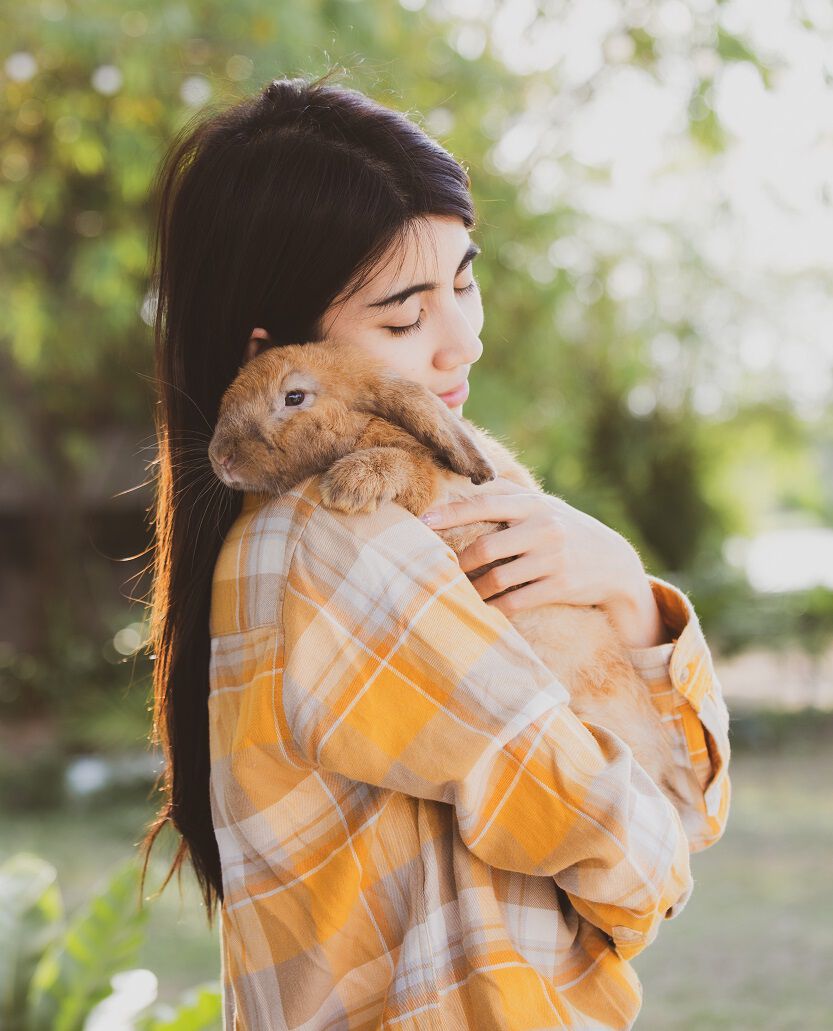A young girl in an orange plaid shirt snuggling with a rabbit outdoors.
